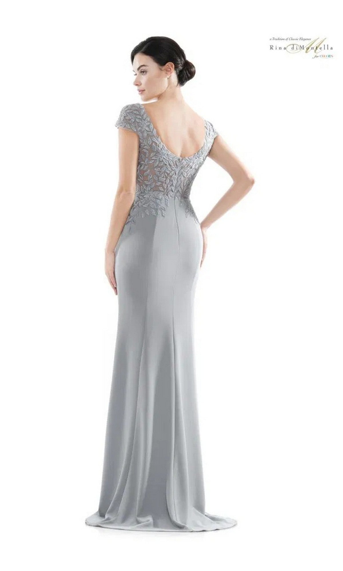 Woman wearing the Rina Di Montella RD2718 long formal dress—silver, floor-length, with an embroidered bodice, lace on the upper back, and cap sleeves—standing against a plain white background.